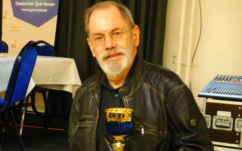 Klaus Otto Nagorsnik mit einem Siegerpokal in den Händen vor dem Wappen des Deutschen Quizvereins